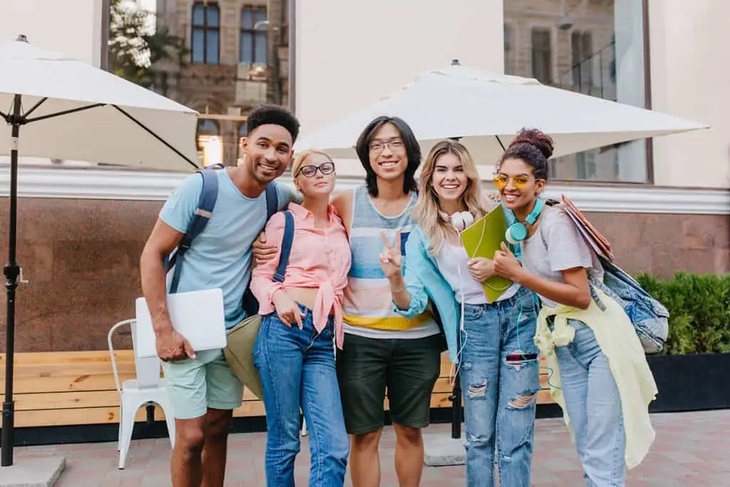 Laughing asian boy in glasses and shorts embracing charming blonde girls in front of outdoor cafe. Joyful students came to open-air restaurant to celebrate end of exams.